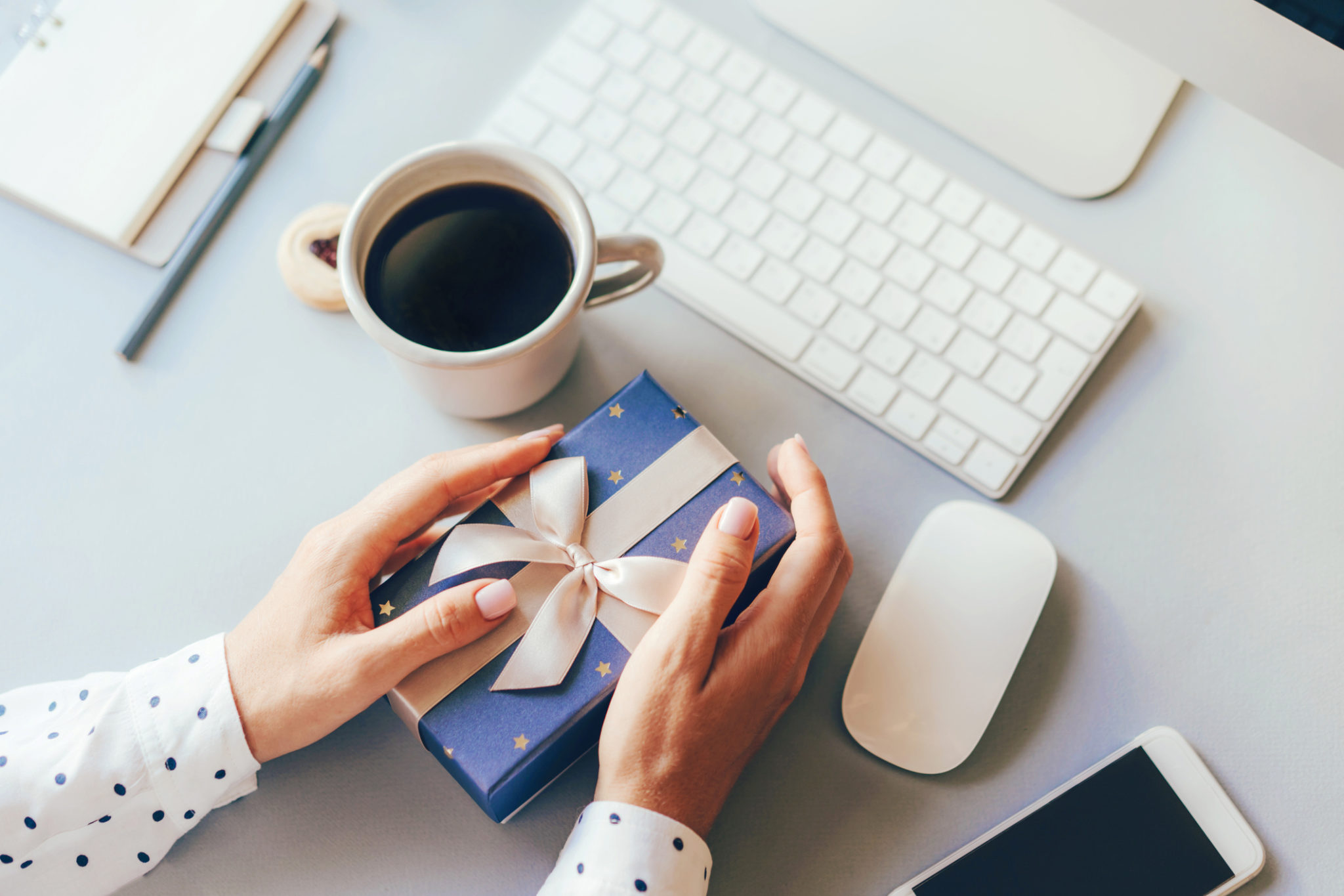 Blue gift box decorated with a golden ribbon bow in female hands against the background of the office workplace, gift delivery, coffee break during office hours, corporate greetings and parties.